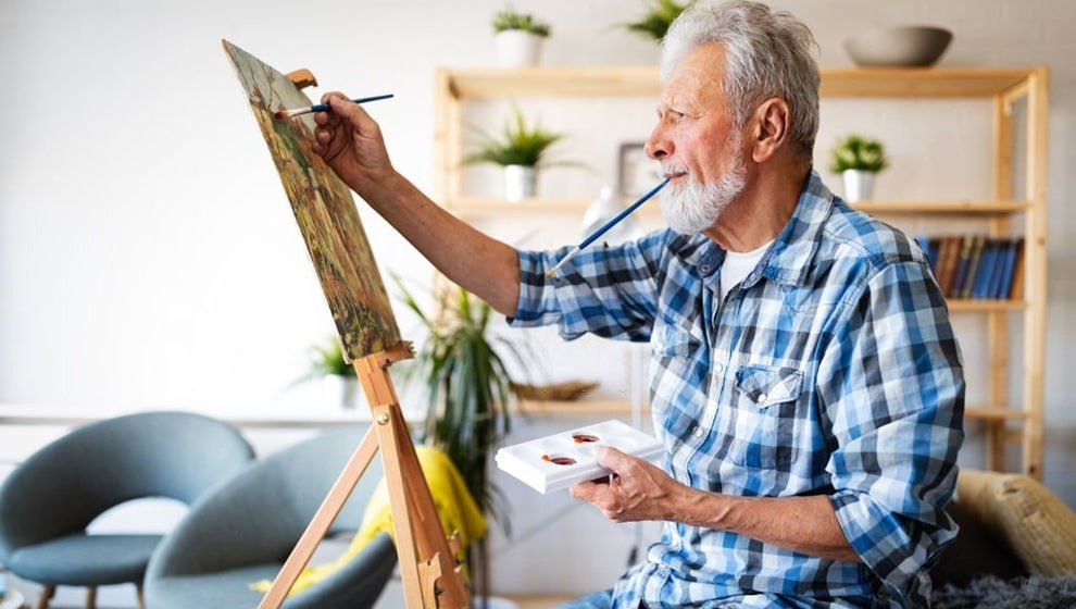An elderly man with a white beard and a blue plaid shirt painting on a canvas attached to a wooden easel at home, holding a palette in one hand and a paintbrush in his mouth while working.