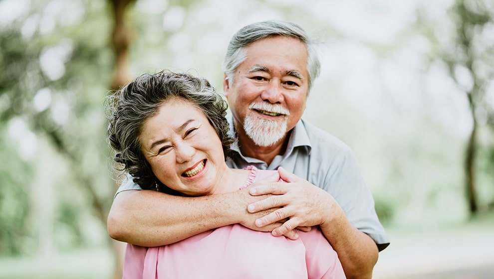 A close-up of a joyful senior East Asian couple laughing together in a park, with the man lovingly embracing the woman from behind against a soft, blurred green background.