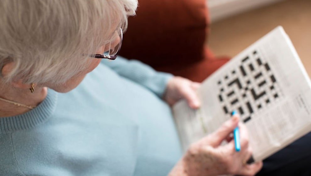 An over-the-shoulder view of a senior woman with short white hair and glasses holding a newspaper or puzzle book, focused on solving a crossword puzzle with a blue pen.