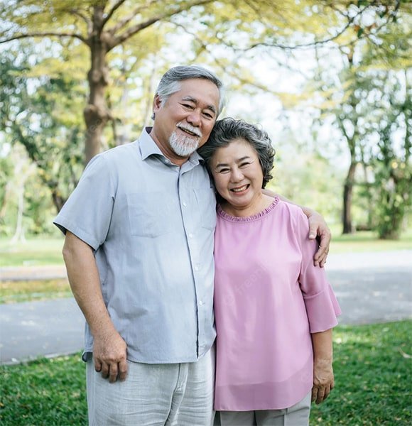 A happy, smiling senior East Asian couple standing close and embracing outdoors in a park with green trees and grass in the background.